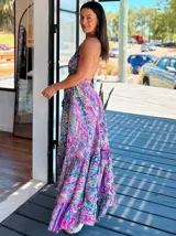 Vestido largo tipo solera, con escote en V y tirantes finos, confeccionado en seda estampada con motivos florales y patrones geométricos en tonos azules, naranjas y morados. Presenta un corte amplio y volados en el ruedo.