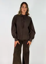 Camisa negra de corte holgado con cuello tipo bebé con volados y lazo de ajuste, cierre frontal con botones y mangas largas abullonadas con puños abotonados.