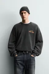 Sudadera de corte relajado, color negro con efecto lavado, cuello redondo y manga larga. Presenta un pequeño estampado frontal con texto en tonos naranja y amarillo.