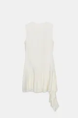 Vestido corto sin mangas, color blanco roto o crema, confeccionado en tejido de encaje. Presenta un bajo con volante asimétrico que cae en cascada hacia un lado.