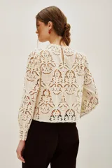 Camisa de encaje negro con cuello alto y mangas largas abullonadas. Presenta un diseño calado con motivos florales y cierre posterior.