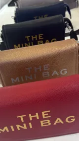 Cartera tipo bandolera de cuero sintético con textura granulada y solapa frontal. Presenta el texto 'THE MINI BAG' estampado en el frente. Incluye correa ajustable y desmontable.