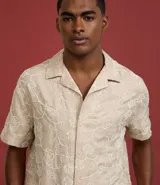 Camisa de poliéster color beige con bordado floral blanco, de corte regular, cuello inglés, manga corta y abotonada en el frente.