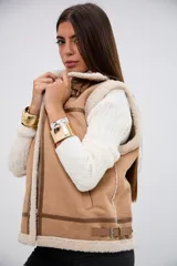 Chaleco de gamuza sintética color camel con interior y bordes de corderito sintético color crema. Presenta cierre frontal metálico, detalles de tiras con hebillas en el cuello y en la parte inferior, y costuras reforzadas con ribetes de cuero sintético.