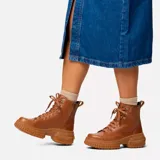 Bota de caña media Sorel, confeccionada en cuero impermeable color marrón tostado. Presenta un diseño moderno con cordones frontales, herrajes metálicos en forma de D y una suela de caucho robusta con plataforma dentada. Incluye tirador en el talón y cierre lateral para mayor practicidad.