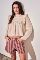 Blusa de color arena con diseño romántico, presenta volados en los hombros, detalles de encaje en el pecho y mangas largas con puños elásticos.
