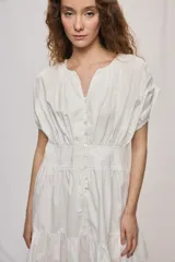 Vestido corto de algodón color blanco, con escote en V y botonadura frontal. Presenta mangas cortas holgadas, cintura ajustada con frunces y falda acampanada con terminación de volado en el ruedo.
