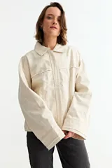 Campera de jean color crudo con diseño de corte holgado, cuello clásico, cierre frontal y detalles de desgastes en toda la prenda.