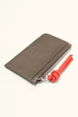 Monedero rectangular de cuero vacuno con textura granulada en color marrón topo. Cuenta con cierre superior metálico y un tirador decorativo de cuero en color naranja con un nudo. Presenta un pequeño logo grabado en el centro.