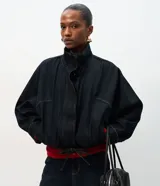 Chaqueta bomber de denim azul oscuro con cuello alto, cierre frontal con zíper y cintura ajustable con cordón. Presenta mangas largas con puños elásticos y bolsillos frontales funcionales.