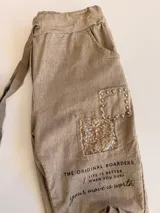 Pantalón de corte relajado color beige, con cintura elástica y cordón ajustable. Presenta parches decorativos con lentejuelas y tachas en los muslos, además de detalles de texto estampado. Posee bolsillos laterales y dobladillo en los tobillos.