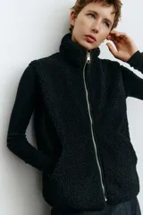 Chaleco de sherpa color negro con cuello elevado y cierre frontal. Presenta ribetes de cinta en los bordes, bolsillos tipo parche en la parte delantera y forro interior.