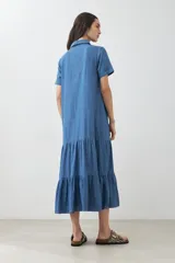 Maxi vestido camisero de jean azul con cuello clásico, manga corta, cierre frontal con botones y falda amplia con volantes.