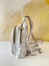 Mochila de cuero blanca con diseño artístico pintado a mano en tonos dorados, marrones y negros en el frente, múltiples bolsillos con cierre y herrajes de metal.