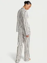 Pijama largo compuesto por una camisa de manga larga con cuello solapa, cierre frontal de botones y bolsillo en el pecho, y un pantalón de corte recto con cintura elástica. Presenta un diseño de rayas verticales oscuras sobre fondo blanco con sutiles detalles florales.