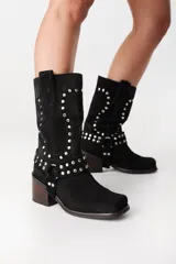 Bota de caña media confeccionada en cuero gamuzado negro, con diseño de estilo western. Presenta detalles de tachas metálicas plateadas en la caña y una correa desmontable con tachas alrededor del tobillo. Cuenta con taco cuadrado de madera y punta cuadrada.