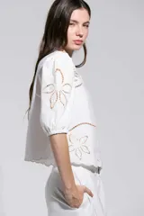 Top Allegra de color blanco, con mangas abullonadas a la altura del codo y puños elásticos. Presenta detalles de calados y bordados florales en los hombros, mangas y ruedo.