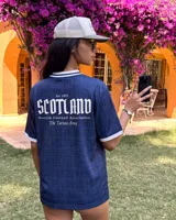 Camiseta de fútbol de Escocia de manga corta, color azul marino con diseño a cuadros sutiles, cuello en V y cuello camisero blanco con líneas azules. Presenta detalles estampados en el frente, incluyendo el escudo de la Asociación de Fútbol de Escocia, un león rampante y el texto 'Scottish Football Association'.
