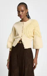 Cardigan de punto color amarillo claro, con cierre frontal de botones metálicos, cuello redondo y mangas largas abullonadas con puños ajustados. Presenta un diseño asimétrico con un panel de punto calado texturizado aplicado sobre uno de los hombros.
