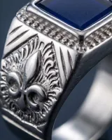 Anillo de acero tipo sello con piedra central rectangular color azul y diseño lateral de flor de lis.