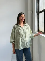 Camisa verde claro de corte oversize, con mangas tres cuartos abullonadas, escote en V con cuello camisero y cierre frontal parcial de botones. Cuenta con delicados bordados florales tono sobre tono distribuidos por toda la prenda.