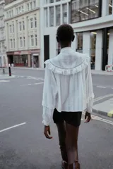 Blusa blanca de manga larga con cuello redondo, confeccionada con detalles de bordados y volantes a tono. Presenta cierre frontal con botones ocultos y puños ajustados.