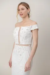 Vestido largo de novia de tul color blanco, con diseño de hombros descubiertos, mangas cortas caídas y corsé con apliques bordados a mano. Presenta una falda sirena con detalles florales bordados y cola.