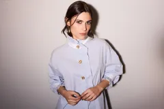 Camisa celeste a rayas verticales azules y marrones, con cuello blanco, cierre frontal con botones dorados y volantes verticales.