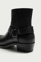 Bota de media caña color negro con detalle de arnés y hebilla lateral, punta perfilada y tacón bajo.