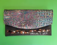Sobre de mano rectangular de Carolina De Cunto, confeccionado en cuero. Presenta una solapa con textura símil cocodrilo en tonos tornasolados y cuerpo inferior negro con detalles de pintura artesanal. Cuenta con cierre de imán y compartimentos internos.