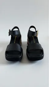 Sandalias de cuero negro Carolina De Cunto, con taco bloque medio intervenido con diseño artístico y hebilla metálica grande en tono oro viejo con logo grabado.