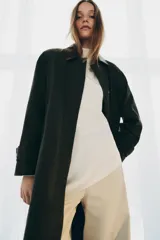 Trench oversize de color verde oscuro, con cuello de piel en tono marrón a contraste. Presenta manga larga con trabillas en los puños, bolsillos delanteros, cierre frontal con cremallera y botones ocultos bajo solapa, y abertura en la espalda.