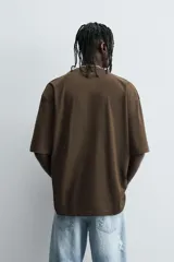 Camiseta de algodón color marrón oscuro, corte relajado (relaxed fit) y manga corta. Presenta un estampado frontal con un lobo o cánido corriendo, el texto "The Riders Project", "VISION & TEMPER" y "0.79 per gallon" en tonos grises y rosados.