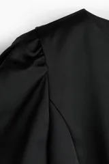 Blusa corta de sarga negra con leve brillo, ajuste entallado, escote redondo, botones forrados al frente y mangas cortas abullonadas con pliegues. Presenta costuras princesa al frente para estilizar la silueta.