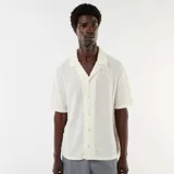 Camisa de manga corta color blanco, confeccionada en tejido de punto calado con patrón de rayas verticales sutiles, cuello abierto tipo campamento y cierre frontal de botones.
