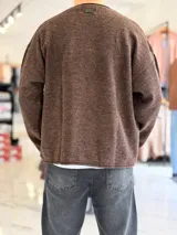 Sweater de punto liviano color beige, con textura marcada, cuello redondo y mangas largas de corte relajado, con pequeño estampado tipográfico negro en el lateral inferior.