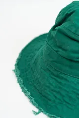Gorro tipo bucket color verde, con ala ancha y bordes deshilachados. Incluye una pequeña hebilla metálica decorativa en el lateral.