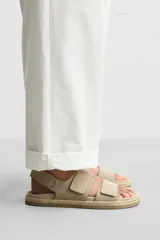 Sandalia color beige de piel con acabado serraje. Cuenta con dos tiras anchas en el empeine y tira en talón ajustables mediante cierres adherentes. Plantilla con horma anatómica y piso forrado de yute.