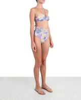 Top de bikini estilo bandeau con estampado floral en tonos violeta y azul sobre fondo crema. Presenta un diseño con nudo central, tirantes finos ajustables al cuello y detalle de cuentas decorativas en la base de los tirantes.