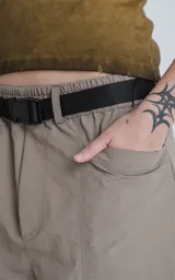 Bermuda técnica color arcilla (marrón claro/beige) de calce oversize, confeccionada en poliéster liviano. Presenta múltiples bolsillos cargo con tapa y un cinturón de ajuste con hebilla plástica negra. Incluye detalles reflectivos.