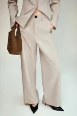 Pantalón de tiro alto en color beige, con diseño de pierna recta y costuras verticales marcadas. Presenta cierre frontal con cremallera y un botón negro en contraste.