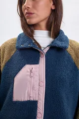 Campera de sherpa con diseño color block, combinando cuerpo azul, mangas marrones y detalles en rosa pálido. Presenta cierre frontal con botones a presión, bolsillo superior con cierre y bolsillos laterales con cremallera.