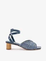 Sandalias de tacón medio en color azul, con capellada de rafia labrada en tonos azules y dorados, pulsera de cuero nobuck enlazada al tobillo con cierre de hebilla, y taco de madera natural lustrada de forma cilíndrica. Cuenta con puntera cuadrada.