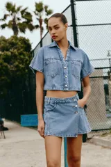 Camisa corta de denim en color azul claro, con cuello solapa, escote en V y manga corta. Presenta bolsillos delanteros de parche con solapa y cierre frontal de botones metálicos.