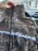Campera de jogging con estampado de camuflaje tipo Real Tree, corte oversize y cierre frontal. Presenta un detalle de puntilla blanca en la parte superior delantera y un bordado a tono en el pecho.