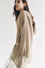 Camisa de cuello fluido y manga larga con puños anchos, de corte recto y largo medio, confeccionada en tejido rústico en tono beige con sutil textura jaspeada. Cuenta con un cinturón ajustable del mismo género para entallar la cintura.