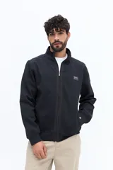 Campera negra de algodón con cierre frontal completo, cuello alto y bolsillos laterales con botón a presión. Presenta un pequeño parche rectangular con texto en el pecho izquierdo.