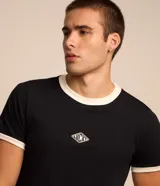 Remera estilo baby tee de color blanco, confeccionada en algodón, con cuello redondo y ribetes en color marrón en cuello y puños. Cuenta con un pequeño tag gomosa con el logotipo "Lucky" en el centro del pecho.