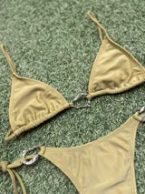 Conjunto de bikini color verde oliva (pistacho) compuesto por un corpiño triangular con argolla central y una bombacha colaless con tiras laterales ajustables y argollas laterales. Las argollas son de zamak.