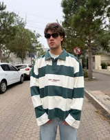 Buzo tipo polo de corte oversize con diseño de rayas horizontales anchas en color gris oscuro y crema. Presenta cuello camisero, cierre frontal parcial con botones ocultos y detalles de costuras expuestas y deshilachadas. Incluye el logo de la marca bordado en el centro del pecho.